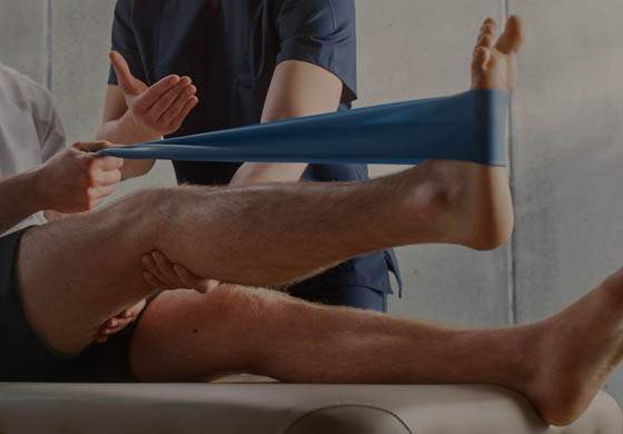 Young man using a resistance band to stretch his leg under the watchful eye of a young, male athletic therapist