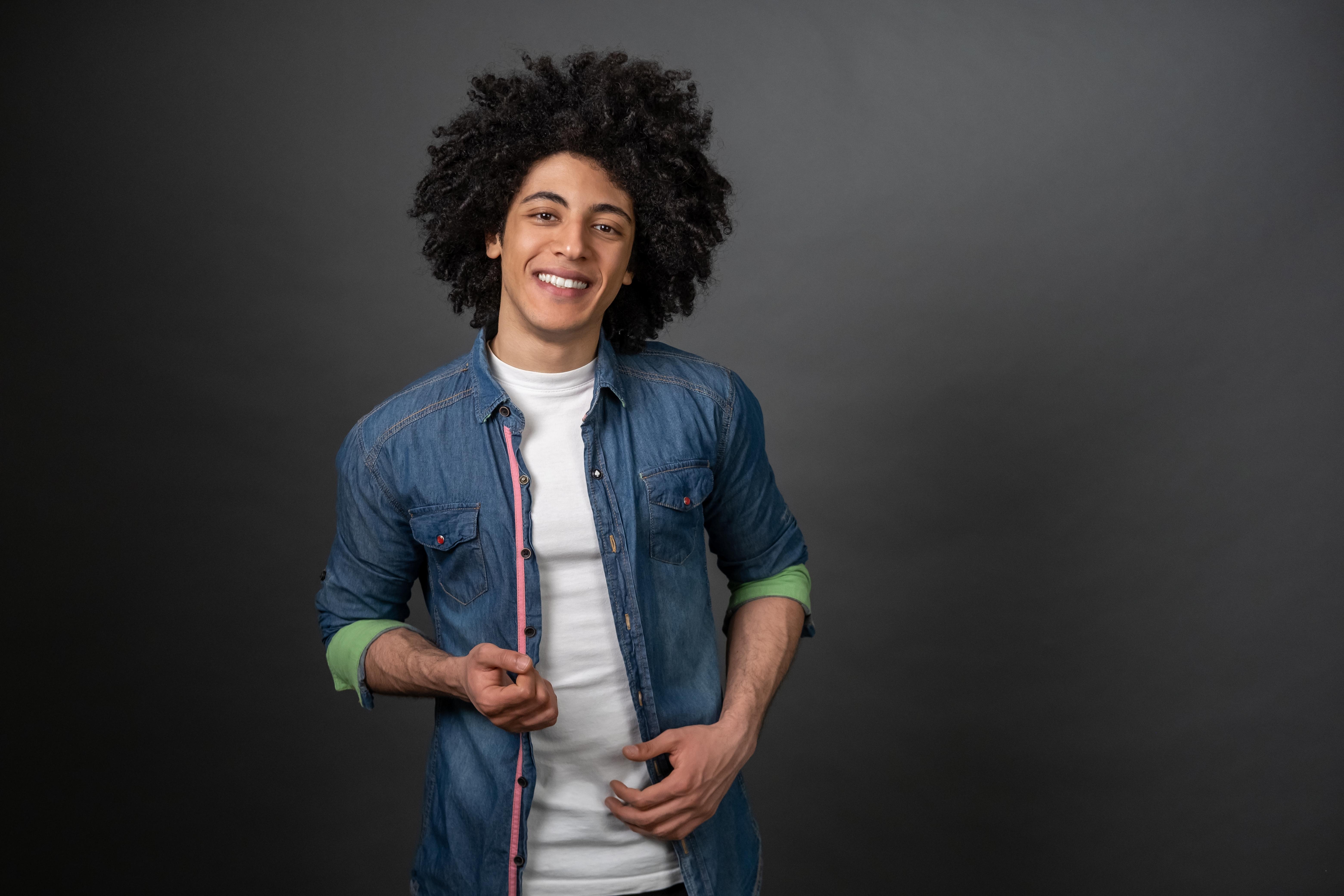 Smiling young man with curly black hair wearing a denim shirt over a white t-shirt against a gray background.